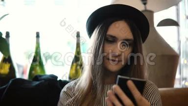 年轻漂亮的女士戴着帽子坐在餐馆里，用智能手机上网，等待点菜