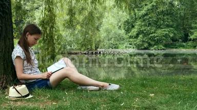女孩坐在树下湖边的公园里看书。
