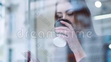 漂亮的年轻女孩穿着时髦的打扮坐在咖啡馆的窗户旁，享受美味的热咖啡，上网，发短信