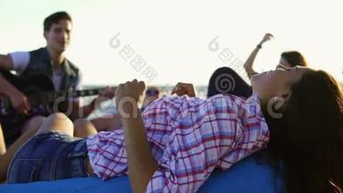 年轻女子躺在安乐椅上，听一个男人在一群坐在海滩上的朋友中间弹吉他