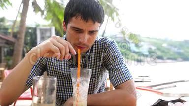 英俊的年轻人坐在海边的海滩咖啡馆里喝鸡尾酒。 慢动作。 3840x2160