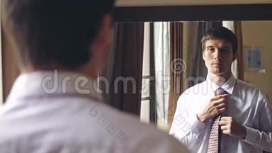 年轻的帅哥穿着白色衬衫站在镜子旁系着领带。 从人到镜子的焦点。 3840x2160