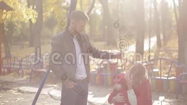 年轻的爸爸妈妈和一个婴儿在操场上玩耍。 公园里为孩子们准备的秋千和旋转木马