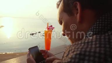 年轻人在日落时使用手机，在露台上喝鸡尾酒，可以看到海景。 慢动作。 1920x1080