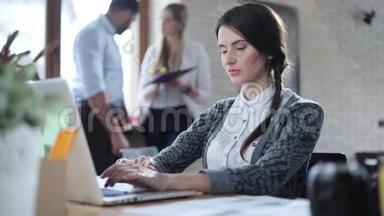 漂亮的黑发女人在轻型办公室的笔记本电脑里打字。 其他工人可以在背景中看到