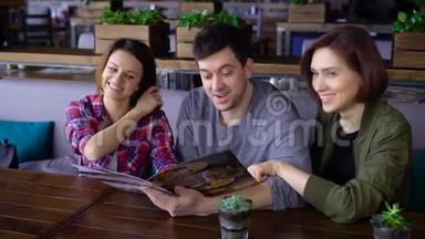 三个快乐的朋友坐在咖啡馆里阅读<strong>菜单</strong>，选择食物和饮料。 <strong>两</strong>个女人和一个男人共进午餐