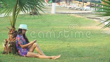 夏天，一个皮肤黝黑的年轻女人，长发坐在热带棕榈树下。 她用智能手机
