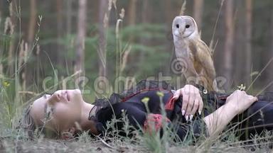 一只长着浅棕色羽毛的可爱的小猫头鹰坐在一个穿着<strong>黑</strong>红色衣服的女孩身上，躺在针叶林里，