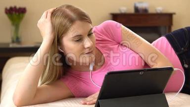 美丽迷人的年轻女子躺在沙发上，在平板电脑上看电影，微笑着。