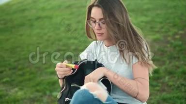 美丽的黑发女孩戴着眼镜，坐在公园的草地上，从背包里拿出<strong>润唇膏</strong>，滋<strong>润</strong>皮肤