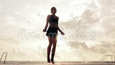 运动的女人穿着短裤和运动胸罩，在海滩边的阳光下，在跳绳上慢动作。 女孩
