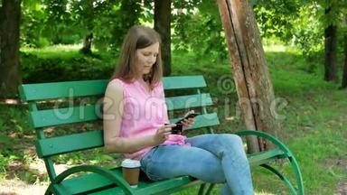 漂亮女孩在公园里坐在长凳上网上买东西。 快乐的女人在网上通过智能手机购物