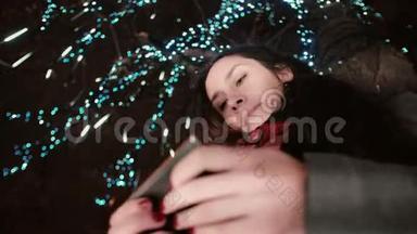 在白雪皑皑的圣诞夜，一个年轻漂亮的女人站在一棵装饰着闪闪发光的树下，用智能手机看着她