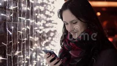 年轻迷人的女人在<strong>圣诞夜</strong>的大雪中使用智能手机站在灯光墙附近，