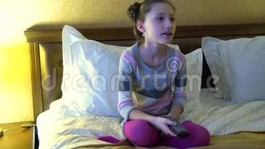 漂亮的小女孩坐在旅馆的床上看电视。 手里拿着遥控器的女孩