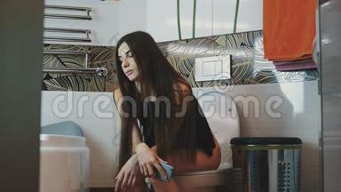 布鲁内特年轻女孩坐在马桶上，手里拿着智能手机。 洗手间。