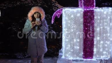 年轻女子正在使用手机，而站在一个巨大的闪闪发光的礼物旁边。