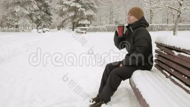 一个年轻人在<strong>冬天</strong>的公园里喝热水瓶里的热饮料。 大雪纷飞。 穿着深色夹克和<strong>保暖</strong>衣服的男人