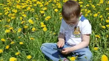 一个快乐的孩子和他的家人在蒲公英的<strong>春天</strong>的小路上。 一个带着<strong>手机</strong>的小男孩坐在绿色的草地上，上面有黄色的