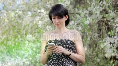 公园里有智能<strong>手机</strong>的年轻女人。 <strong>春天</strong>。 在一棵开花的树的背景下