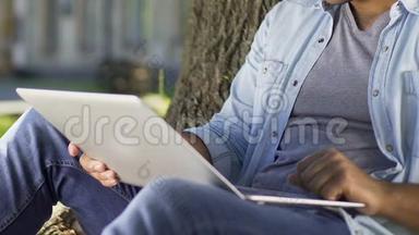 学生在他的笔记本电脑上写报告，坐在大学校园的树下