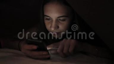 年轻女孩躲在羽绒被下，在黑暗中使用PC平板设备。 青少年晚上在数字平板电脑上玩。