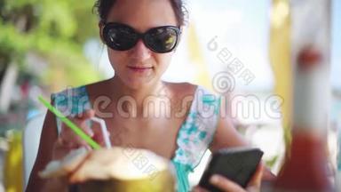 在海滩上，年轻漂亮的女人戴着太阳镜，用手机，喝着椰子做的鸡尾酒，放松心情