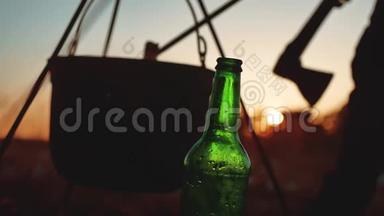 点燃一个大锅篝火，一瓶啤酒在日落剪影露营特写。 旅游、户外休闲、生活方式