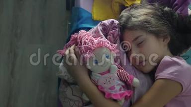 可爱的小女孩晚上睡在室内的沙发床上，拥抱她的娃娃。 女孩深肤色的少年可爱的睡着了