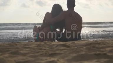 年轻的浪漫夫妇正坐在海滩上拥抱着美丽的景色。 一个女人和一个男人坐在一起