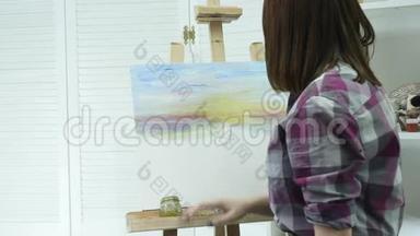 一位年轻女子站在画架后面的一张画布上，在一个大气车间里画一幅画