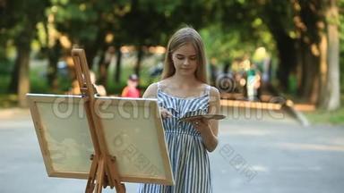 美丽的女孩在公园里画一幅画，用颜料和铲子调色板。 带有图片的易拉罐和帆布.. 夏天是