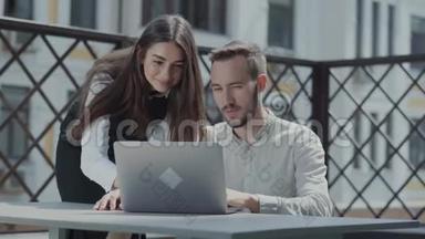 大胡子的年轻人坐在笔记本电脑前的露台上的桌子旁工作。 一个漂亮的女孩弯<strong>下腰</strong>看着