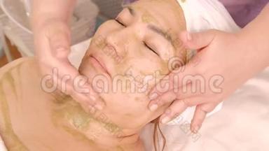 在美容院闭着眼睛的亚洲女孩。 面部按摩用藻类或水果面膜。 专业人士的双手