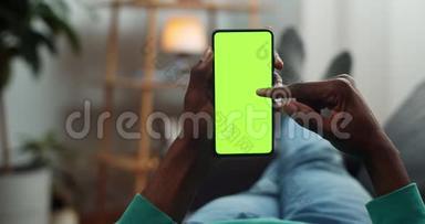 非洲男人滚动智能手机模拟显示时，躺在沙发上。 年轻人一边上网一边看绿色