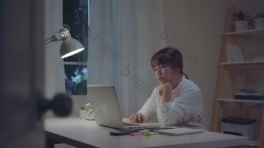 年轻的亚洲妇女在家里的客厅里的桌子上工作到很晚。亚洲女商人写笔记本文件财务和计算器在夜间在家里的办公室。享受在家的时间概念.
