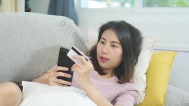 年轻、面带微笑的亚洲女人在家里客厅里躺在沙发上，用智能手机用信用卡上网购物。生活方式，拉丁裔和他的恐慌裔妇女在家的观念.