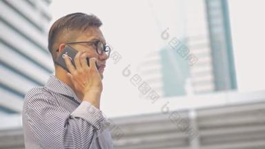 慢动作肖像亚洲商人接电话。一个戴眼镜的亚洲男子在大办公楼附近的街道上用手机.