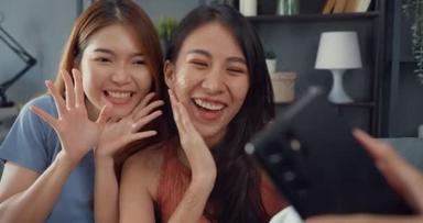 年轻的亚洲女人开心地笑着放松，在家里客厅里使用智能手机视频通话。快乐的室友女士与朋友和家人的视频会议，家庭妇女的生活方式概念.