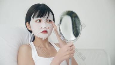 漂亮的年轻亚洲女人，戴着薄片面膜，在家里照镜子，漂亮的女孩用化妆品和化妆品，用皱纹护肤、护肤和保湿霜触摸脸.