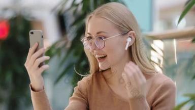 兴奋的年轻迷人的千年女孩，带着无线耳机聊天，通过<strong>视频</strong>通话使用手机应用程序，带着眼镜的高加索女人快乐地笑着虚拟迎接远程通信现代技术