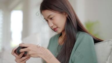 快乐的年轻亚洲女人在家里舒服的沙发上放松，在智能手机上玩网络游戏，微笑的女孩用手机聊天，在电子产品上浏览无线网络，从家里上<strong>网购</strong>物