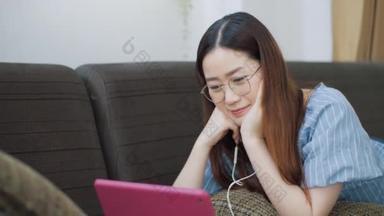 快乐的亚洲女人带着耳机，一边在平板电脑上看电影，一边在沙发上享受着放松的生活.