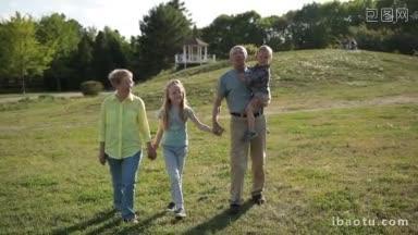迷人的爷爷奶奶带着孙子孙女在公园散步，快乐的爷爷抱着他可爱的刚学步的孙子