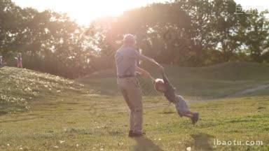 深情的爷爷玩耍和旋转他可爱的孙子，而家人在公园享受休闲在惊人的日落的光辉