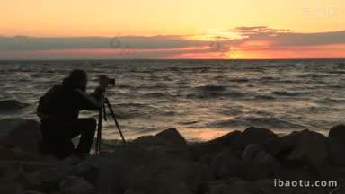 年轻时尚的摄影师与背包制作照片的海洋和岩石与相机在三脚架上的黄昏时间专业