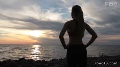 自信的运动苗条的女人站在岩石海岸，享受海景<strong>和</strong>彩色的天空与云在<strong>日</strong>落后的看法