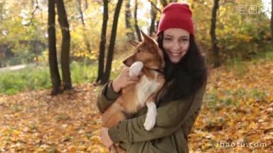 快乐积极的年轻女子穿着派克大衣，戴着红色针织帽，抱着她可爱的狗，对着金色的秋天风景微笑