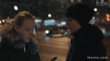 两个女人朋友在城市的街道上，在晚上，她们在聊天，一个女人在用智能手机