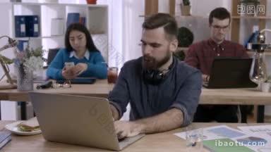 留着胡子的时髦商人坐在办公室的笔记本电脑前吃处方药，而同事们在后台工作